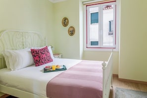 Ein Bett mit weißer Bettwäsche und einer rosa Decke, ein Nachttisch mit einem Tablett Orangensaft und einem Buch, und ein Fenster mit rotem Rahmen.