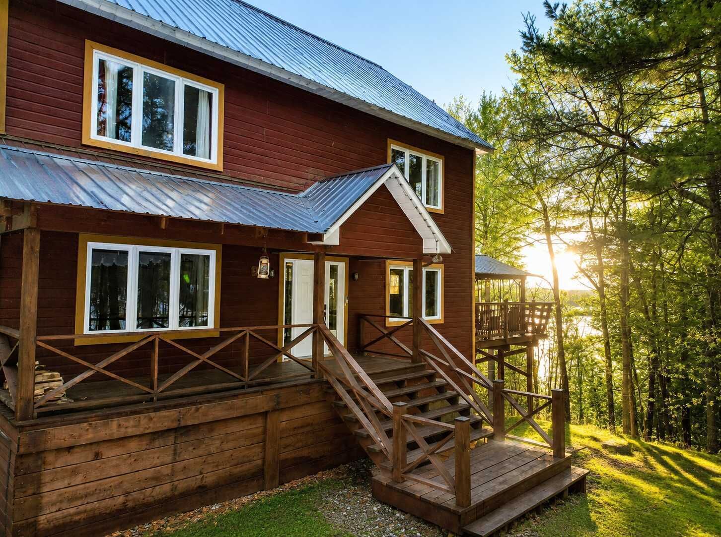 A two-story wooden cabin with a metal roof, a deck with stairs, and a view of a forested area.