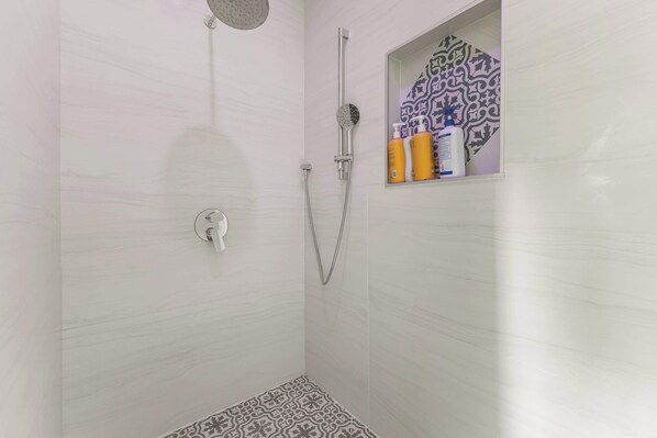 A shower with a handheld showerhead, a wall-mounted faucet, and a small shelf with toiletries.