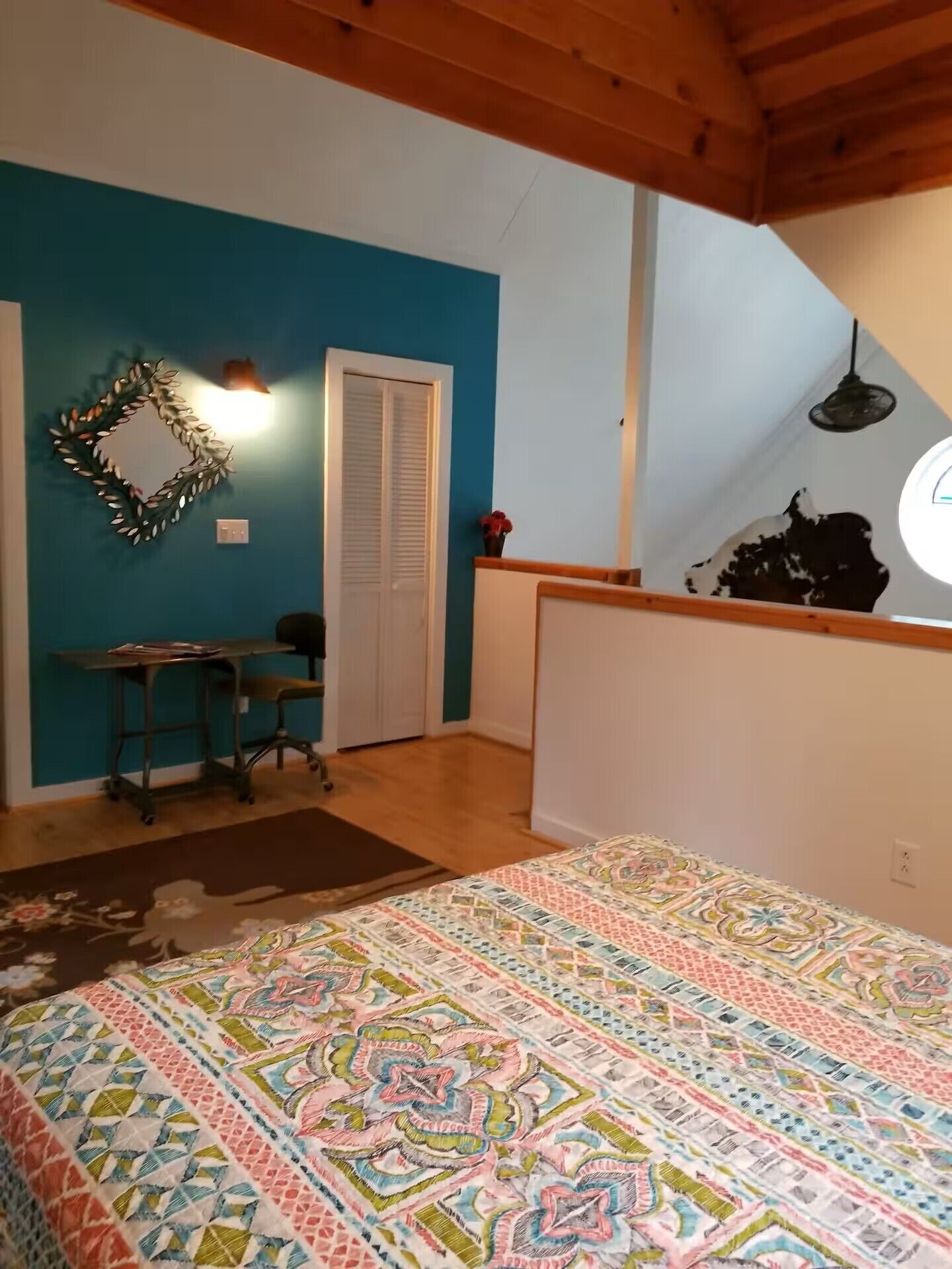 A bedroom with a patterned bedspread, a desk with a chair, a wardrobe, and a mirror on the wall.