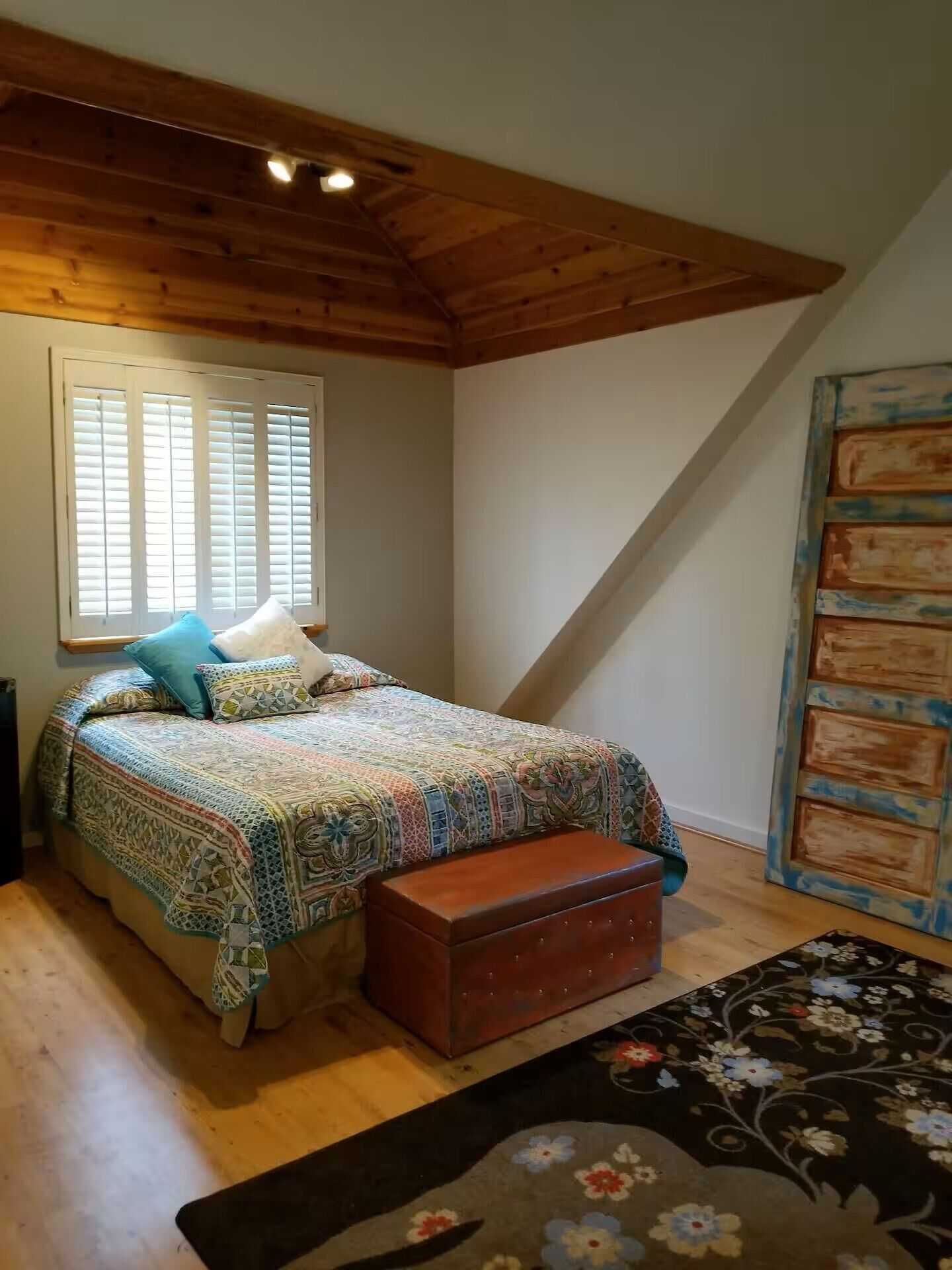 A bedroom with a wooden ceiling, a bed with patterned bedding, a bench, and a large window with blinds.