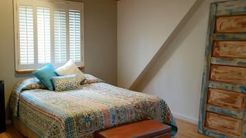A bedroom with a wooden ceiling, a bed with patterned bedding, a bench, and a large window with blinds.