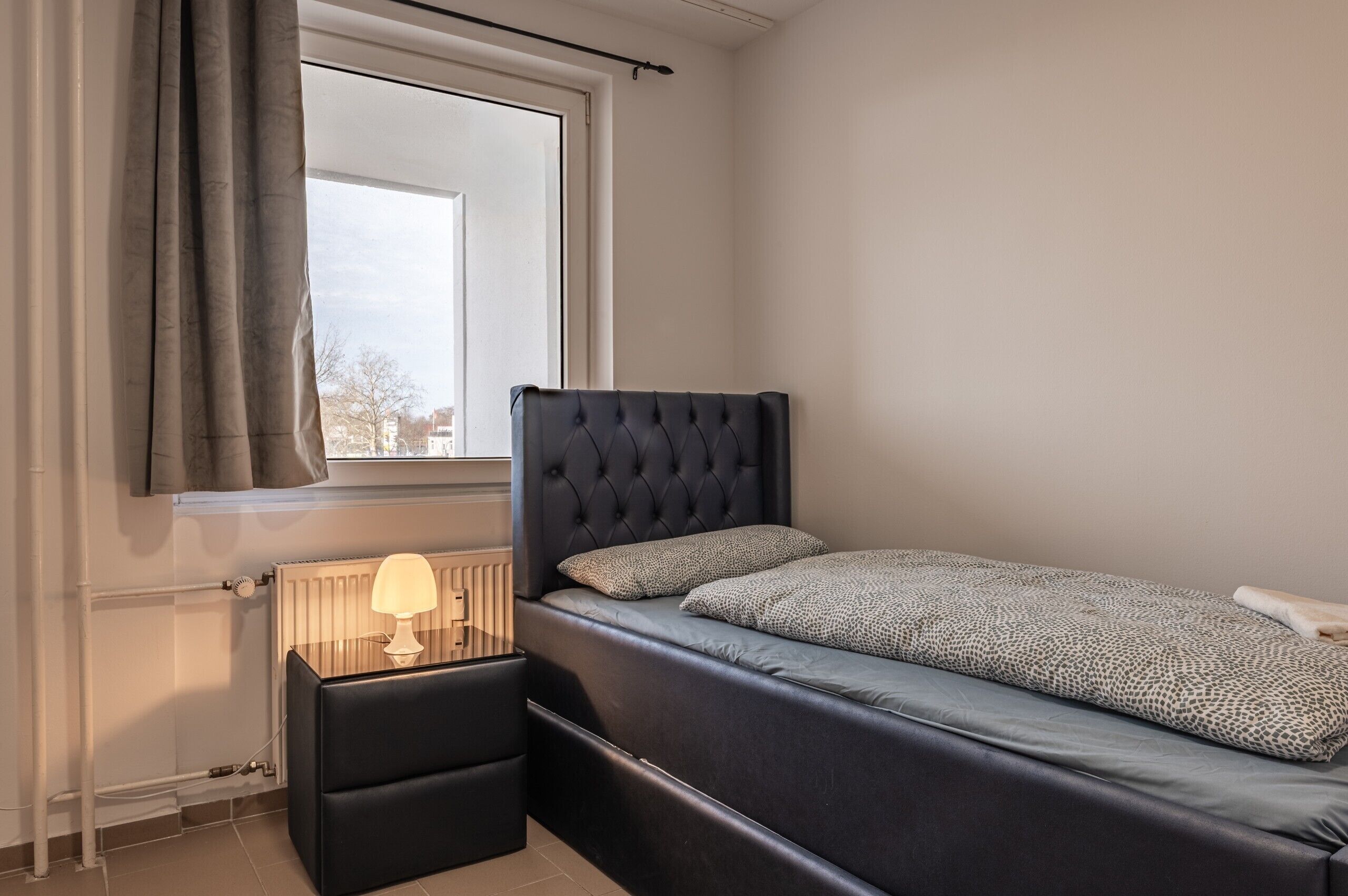 A bedroom with a bed, a bedside table with a lamp, a window with curtains, and a radiator.