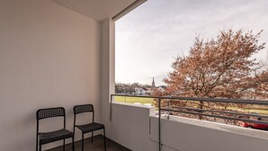 Un balcon avec deux chaises, une balustrade et une vue sur un arbre et des bâtiments éloignés.