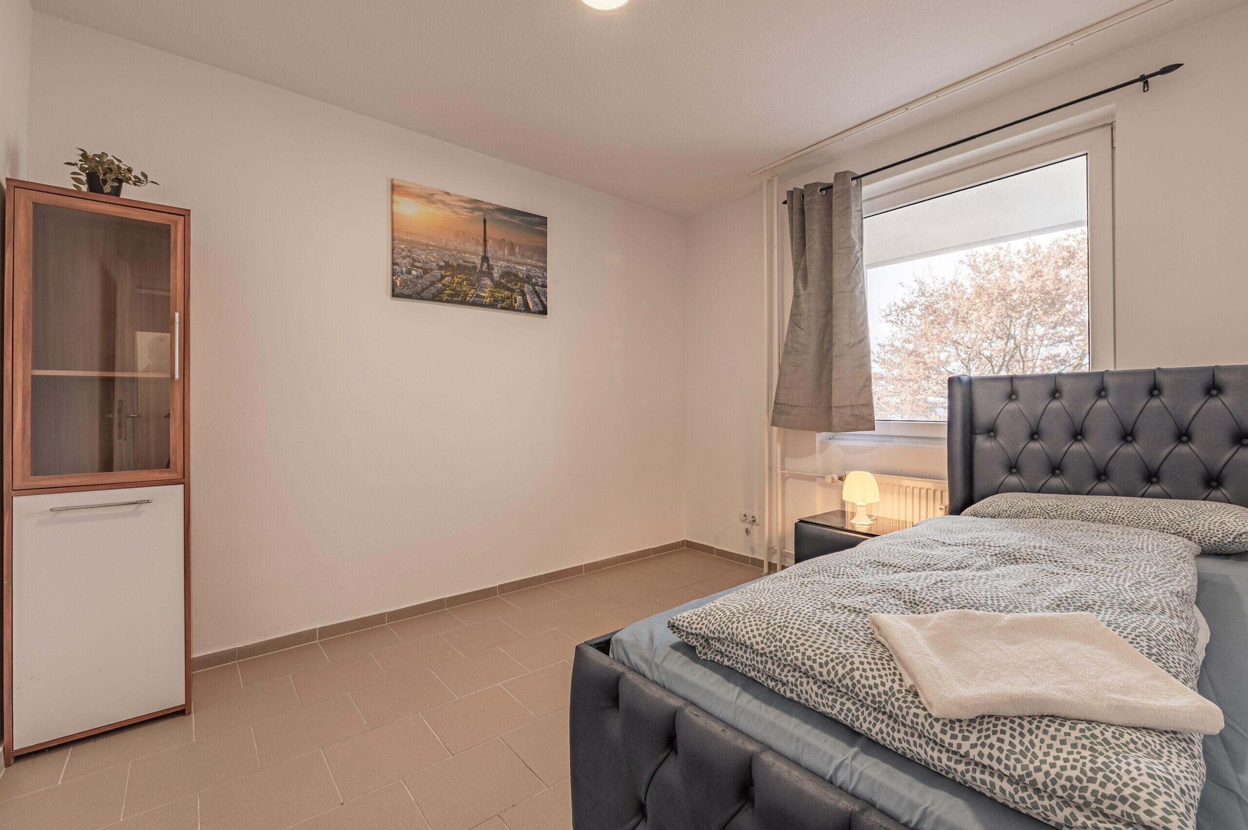 Une chambre à coucher comprenant un lit, une table de chevet, une armoire, une fenêtre avec des rideaux et un tableau représentant un paysage urbain accroché au mur.