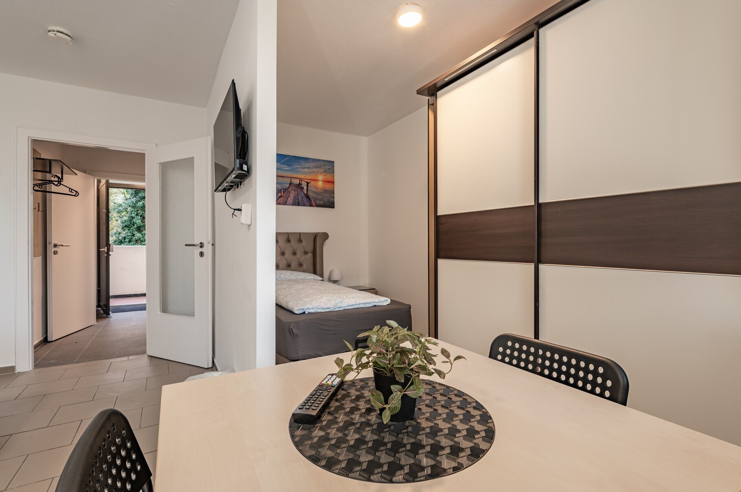 Una habitación de hotel con cama, televisor montado en la pared, una mesa de comedor con sillas y una planta en maceta.