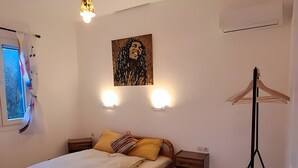 A bedroom with a bed, bedside table, nightstand, wall-mounted light, air conditioning unit, and a window with curtains.
