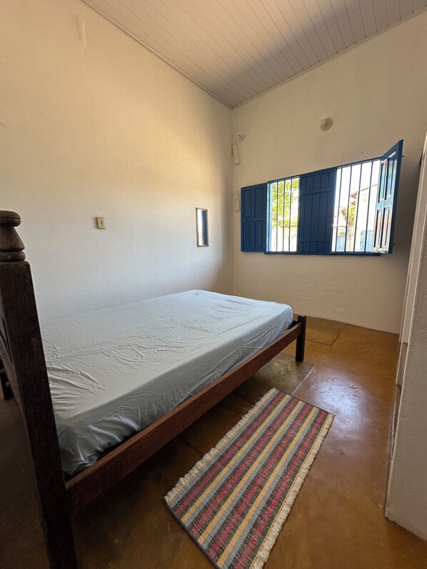 A single bed with a wooden frame and a white mattress, a small rug, and a window with blue shutters.