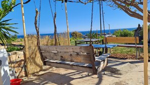 Un banc en bois offrant une vue sur la mer, une table et des chaises, le tout installé dans un espace extérieur ensoleillé.