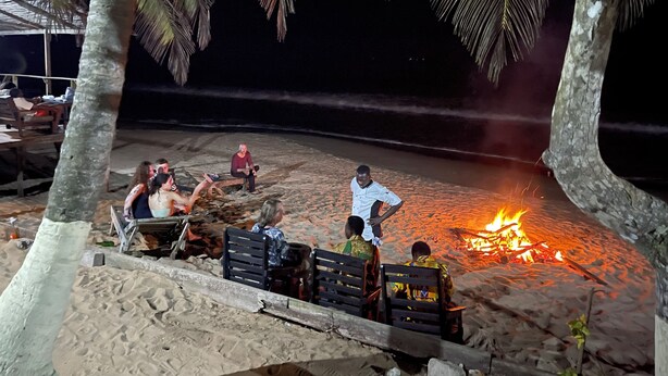 Cena de praia com pessoas sentadas em bancos e cadeiras ao redor de uma fogueira.