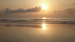 Uma praia com o sol refletindo na areia molhada, ondas se aproximando da costa e um céu nublado.