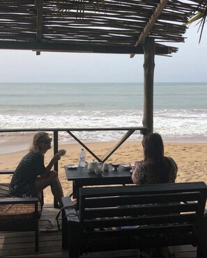 A beachside table with two people seated, a thatched roof overhead, and a view of the ocean. - Simple and Eco friendly Beach Resort and bungalows , seafood ,vegetarian 7 (Takoradi)
