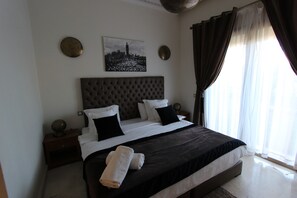 A neatly made bed with a dark headboard, white bedding, and two black pillows. There is a round wall decoration above the bed and a picture of a cityscape on the wall. - Exceptional villa with private pool in Marrakech, Palmeraie area, Atlas view (Marrakech)