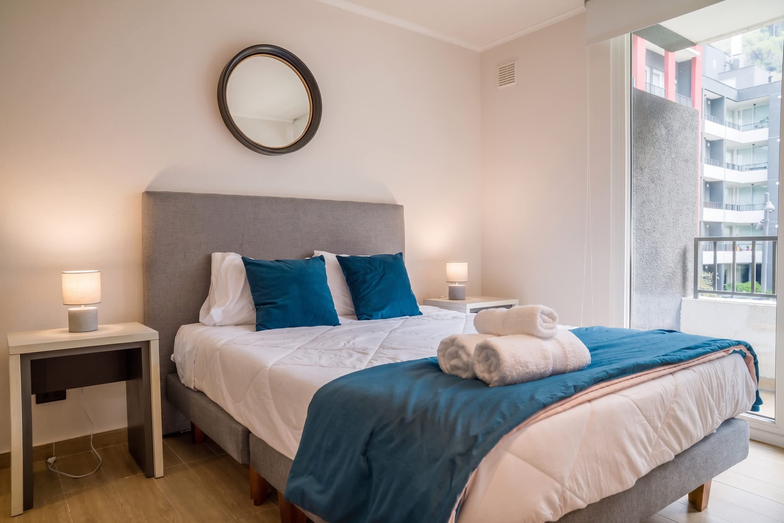 A hotel room with a bed, two bedside tables, a mirror, and a window with a view of buildings.