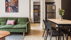 Ein modernes Wohnzimmer mit einem grünen Sofa, einem Esstisch mit Stühlen, einer Küchenzeile mit Kühlschrank und einem Flur zu einem Kleiderschrank.