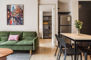 Un salon moderne avec un canapé vert, une table à manger avec des chaises, un coin cuisine équipé d’un réfrigérateur et un couloir menant à un placard.