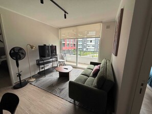 Ein modernes Wohnzimmer mit einer Couch, einem Fernseher, einem Ventilator und einem Balkon mit Blick auf Gebäude.