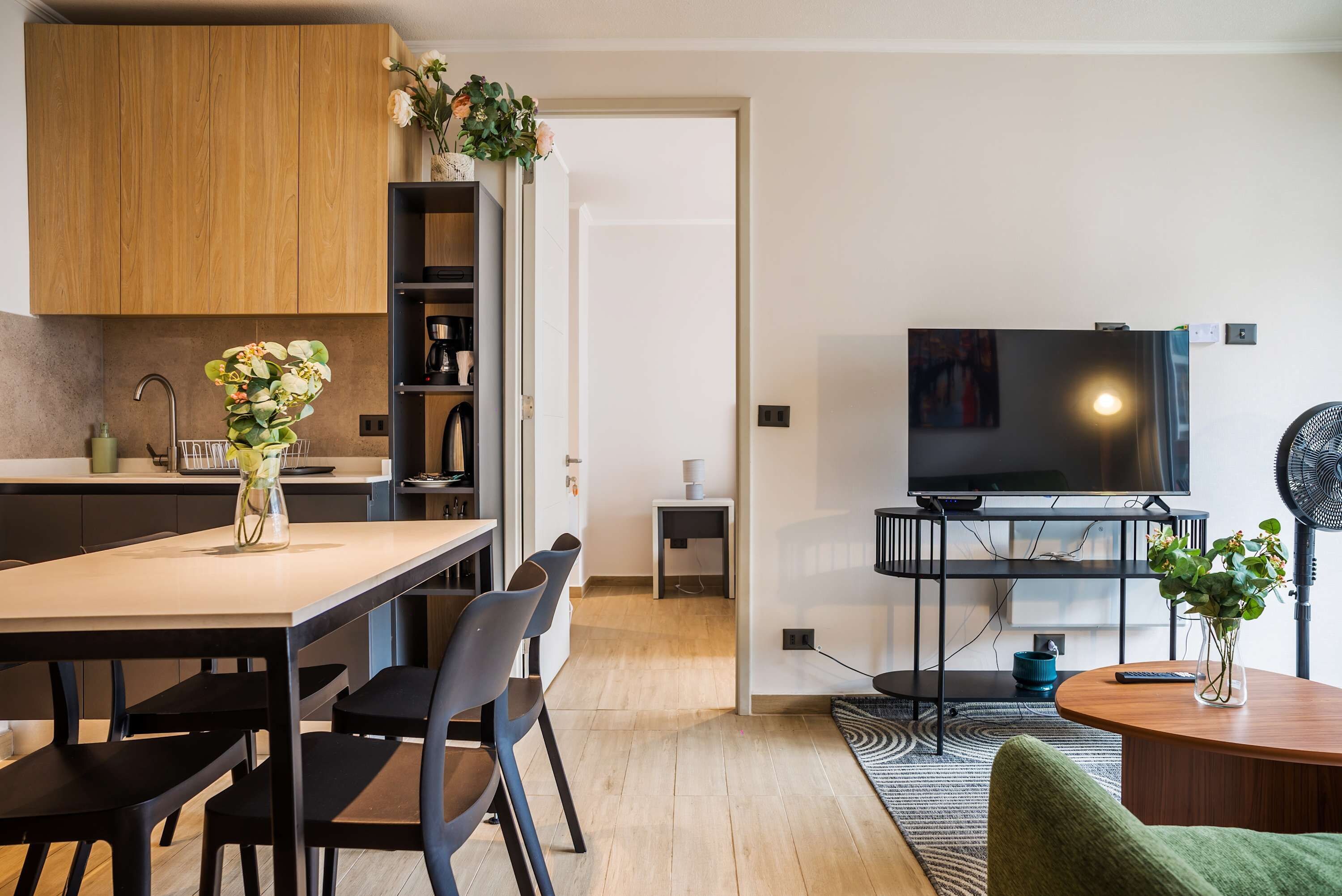 Une cuisine moderne avec des meubles en bois, une table à manger avec des chaises, un téléviseur posé sur un meuble et une petite table sur laquelle se trouve un vase de fleurs.
