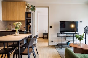 Une cuisine moderne avec des meubles en bois, une table à manger avec des chaises, un téléviseur posé sur un meuble et une petite table sur laquelle se trouve un vase de fleurs.