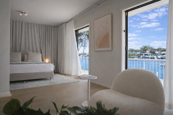 A bedroom with a bed, a chair, a small table, and a large window with a view of a body of water and buildings.