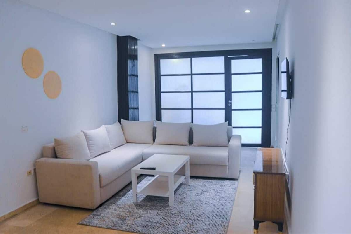 Uma sala de estar moderna com um sofá seccional bege, uma mesa de centro branca e uma pequena mesa lateral de madeira.