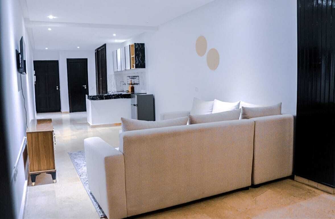 A modern living room with a beige sofa, a wooden side table, and a kitchen in the background.