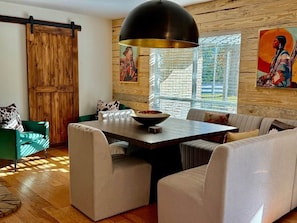 Un salón moderno con mesa de comedor, sofás, una lámpara colgante negra grande, paredes de madera y obras de arte.