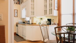 Una cocina con armarios blancos, lavavajillas, cafetera y comedor con mesa y sillas de vidrio.