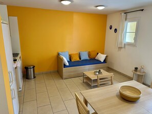A compact living space with a sofa against a yellow wall, a small table, and a window with curtains. - PMR 2/4 persons - Air conditioning - swimming pool - 100m from the beach (Sari-Solenzara)