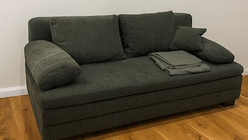 Tummanvihreä sohva, jossa on kaksi koristetyynyä ja taitettu viltti puulattialla.