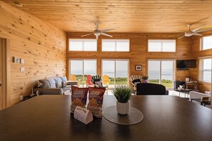Un intérieur en bois avec une grande table, des chaises, un canapé, des ventilateurs de plafond et de grandes fenêtres.