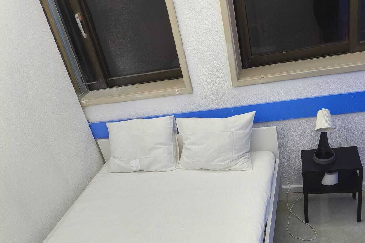 A single bed with white bedding, a nightstand with a lamp, and a window with blinds.