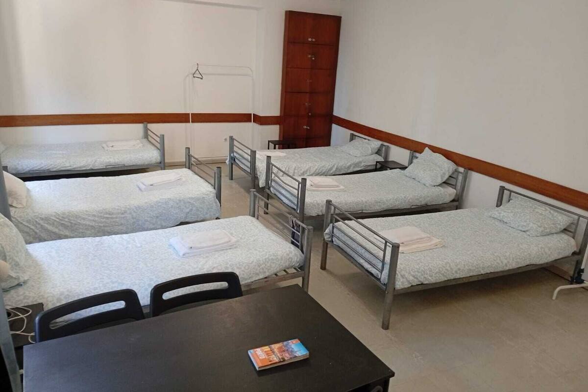 A dormitory room with four single beds, a wooden wardrobe, a table with a magazine, and a chair.