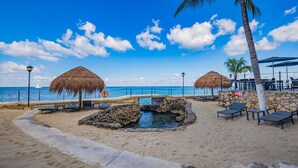 Un complexe hôtelier situé en bord de mer, doté de parasols en chaume, de chaises longues et d’une petite piscine.