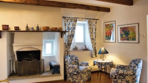 Un salon chaleureux avec une cheminée, deux fauteuils à motifs, une petite table avec une lampe et une étagère ornée d’objets décoratifs.