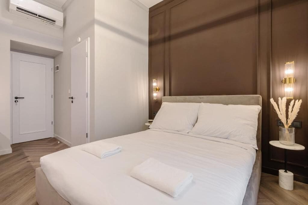 A neatly made bed with white linens and a folded towel, a bedside table with a lamp and decorative flowers, and a door with a handle.