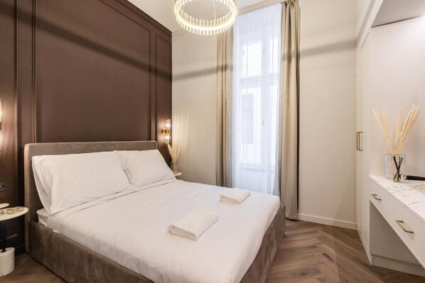 A neatly made bed with white linens and two folded towels, a bedside table with a lamp, and a large window with curtains. - Arany Boutique Residence by BQA (Budapest)