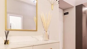 Ein modernes Badezimmer mit einem weißen Waschtisch, einem spiegel mit goldener Rahmen und einem beigefarbenen Sessel.