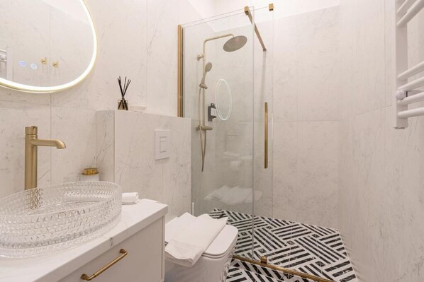 A modern bathroom with a glass-enclosed shower, a white vanity with a gold faucet, and a patterned floor. - Arany Boutique Residence by BQA (Budapest)