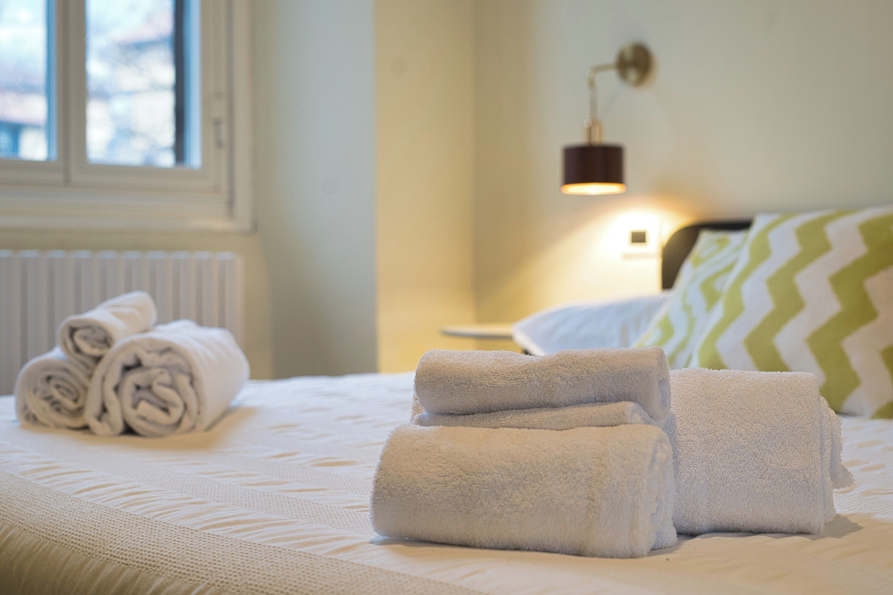 A neatly made bed with rolled white towels, a bedside lamp, and a window with blinds.