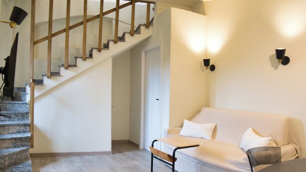 Eine Treppe mit Holzhandläufen führt zu einer weißen Tür, ein beiges Sofa mit Kissen und zwei Wandleuchten.