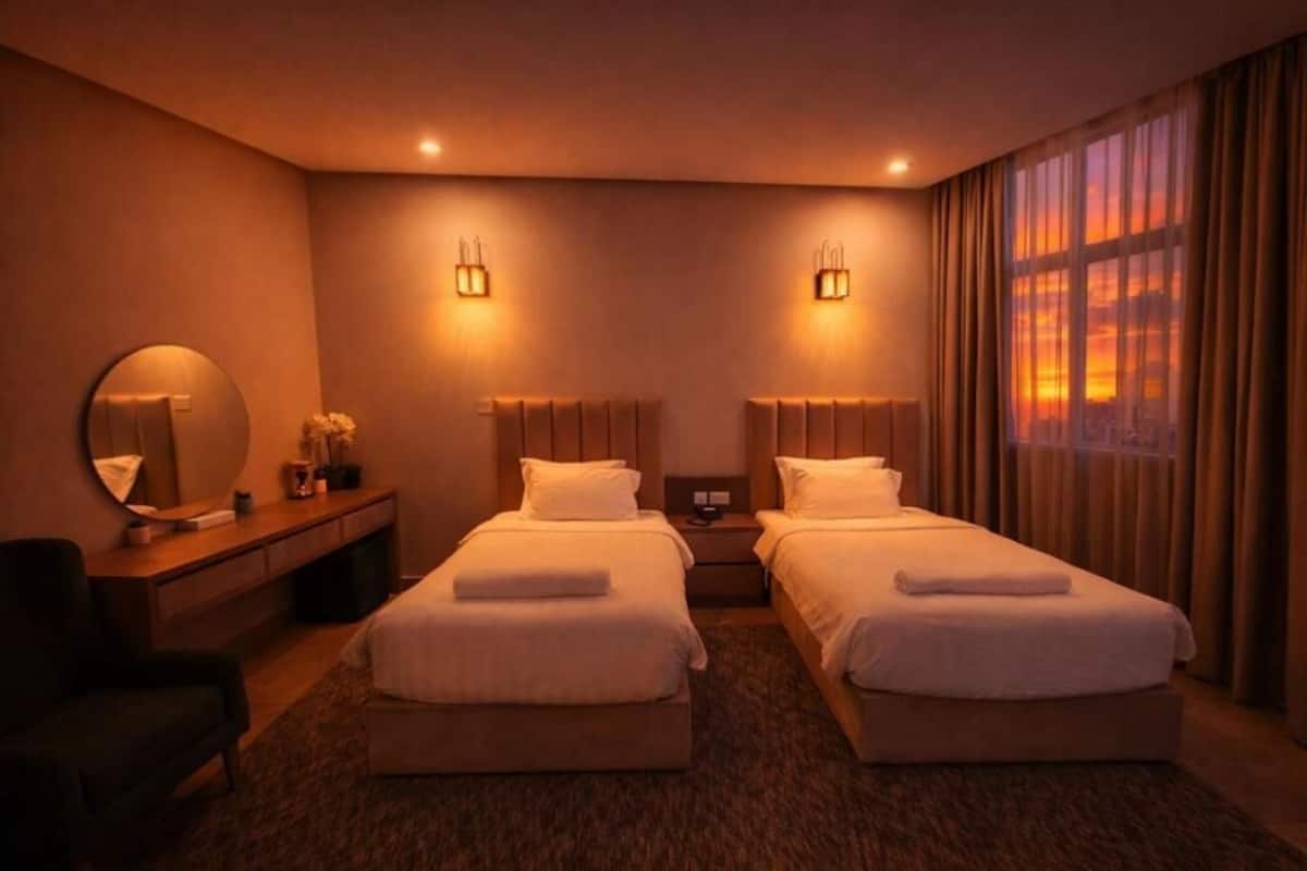 Ein Hotelzimmer mit zwei Betten, einem Schreibtisch, einem Stuhl, einem runden Spiegel und Blick auf den Sonnenuntergang durch das Fenster.