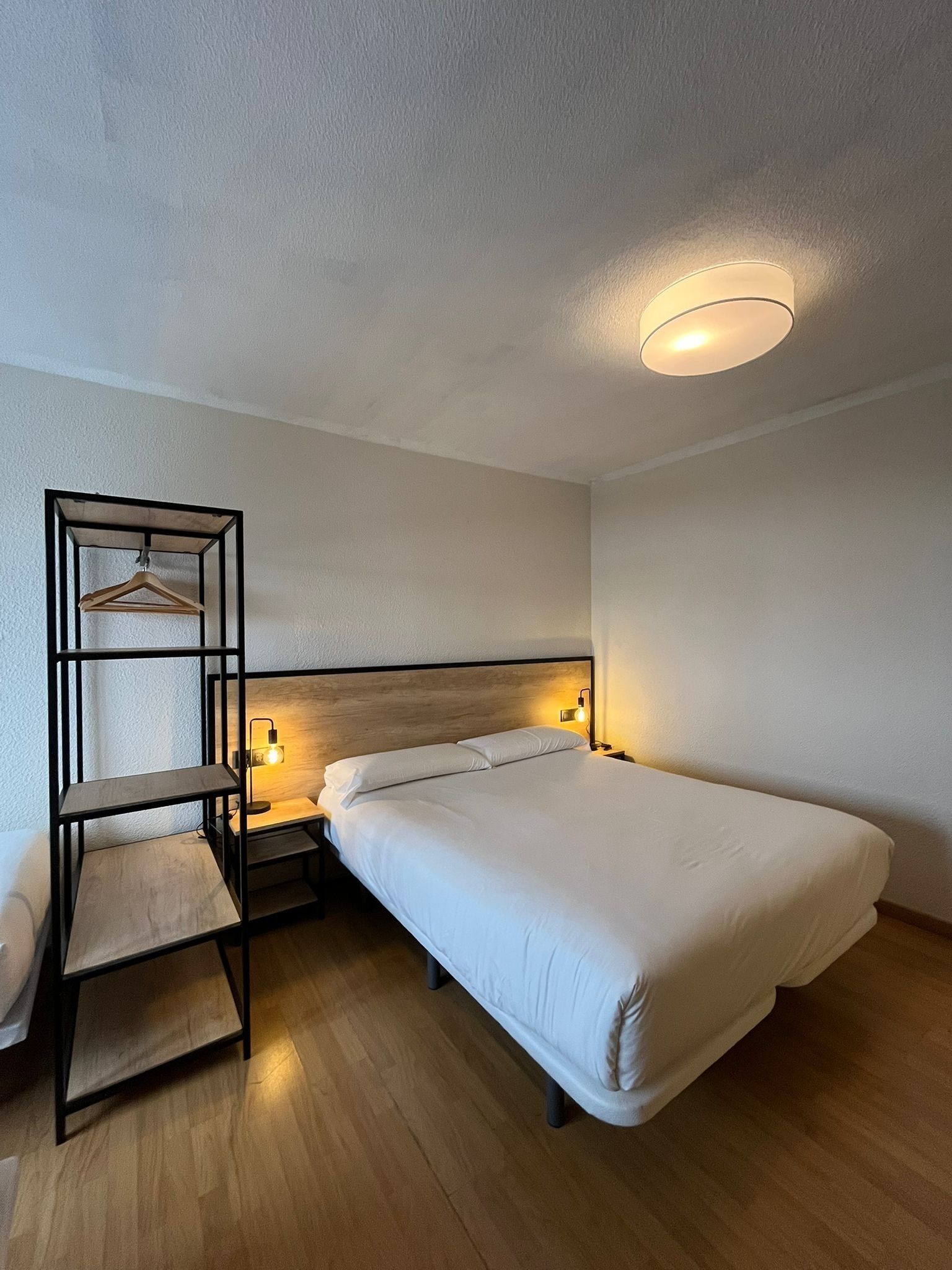 A modern hotel room with a wooden floor, a bed with white linens, a bedside table with a lamp, and a hanging wardrobe.