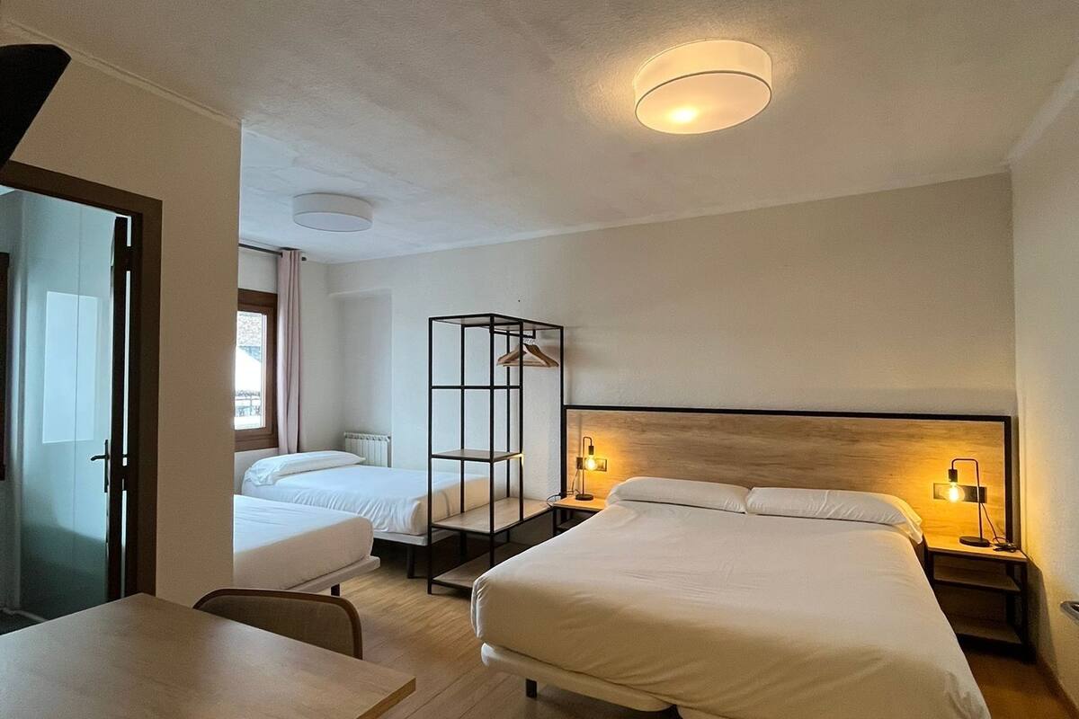 Una habitación de hotel con una cama grande, una cama más pequeña, un escritorio y una silla. Se observa un armario con espejo y estante.