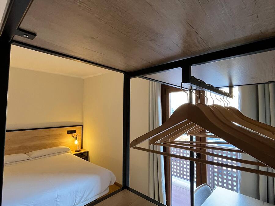 Habitación de hotel con cama, mesita de noche, lámpara y vistas al exterior a través de una ventana.