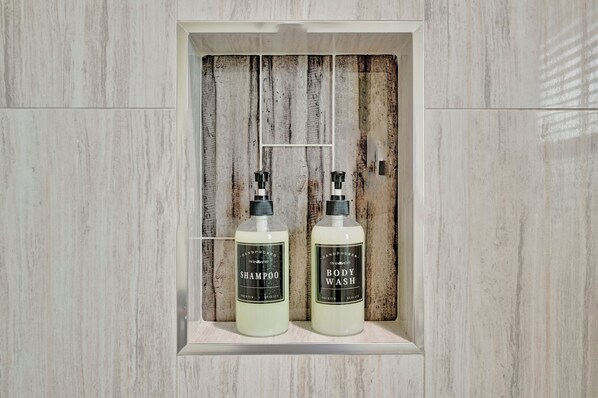 Baño con pared de paneles de madera y dos botellas de champú y gel de ducha artesanales en un mueble blanco.