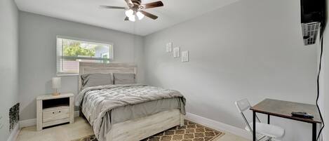 A bedroom with a bed, a desk, a chair, a ceiling fan, a nightstand, and a window.