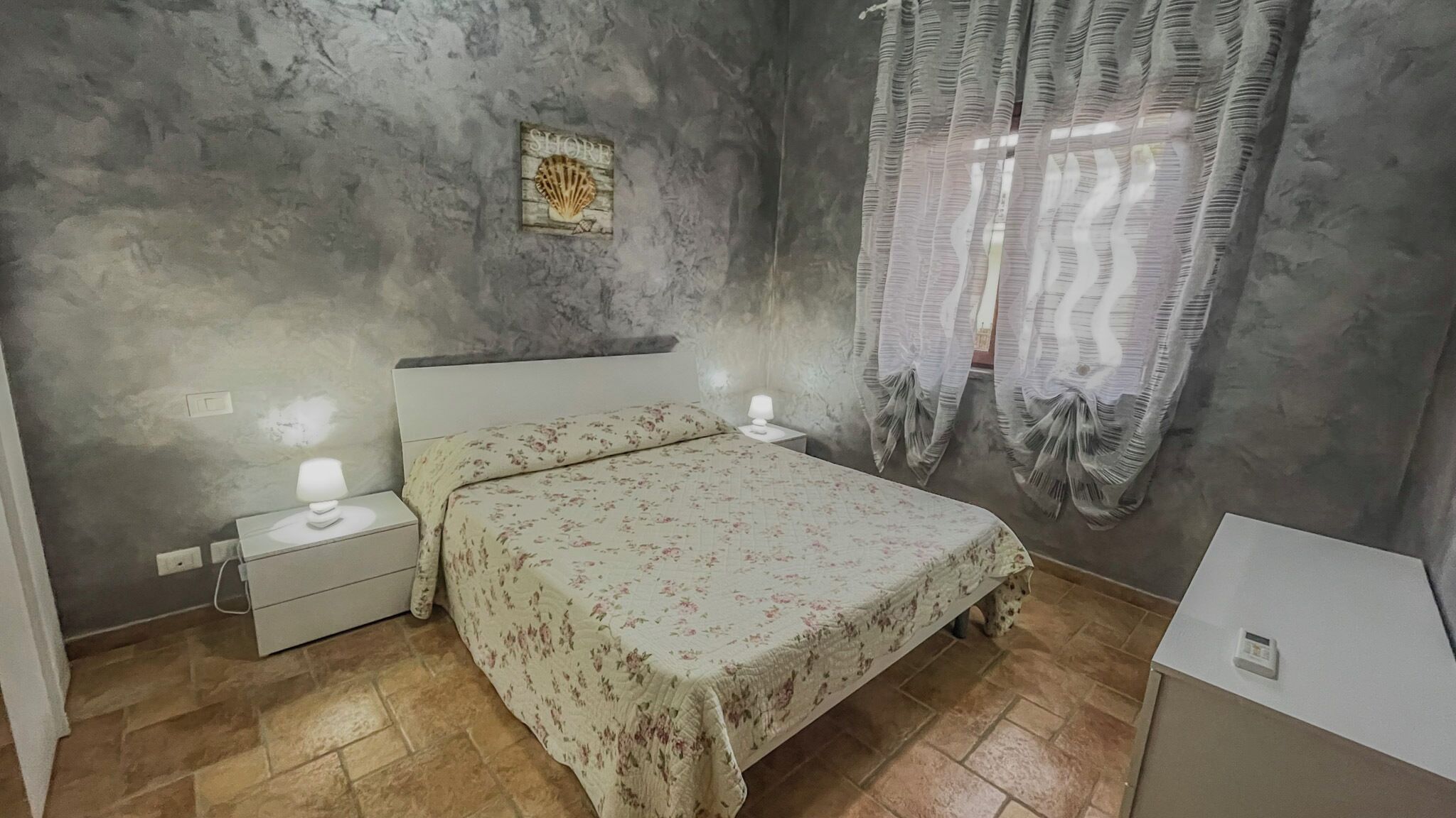 A bedroom with a bed, nightstands, a wall-mounted shelf, and a window with curtains.