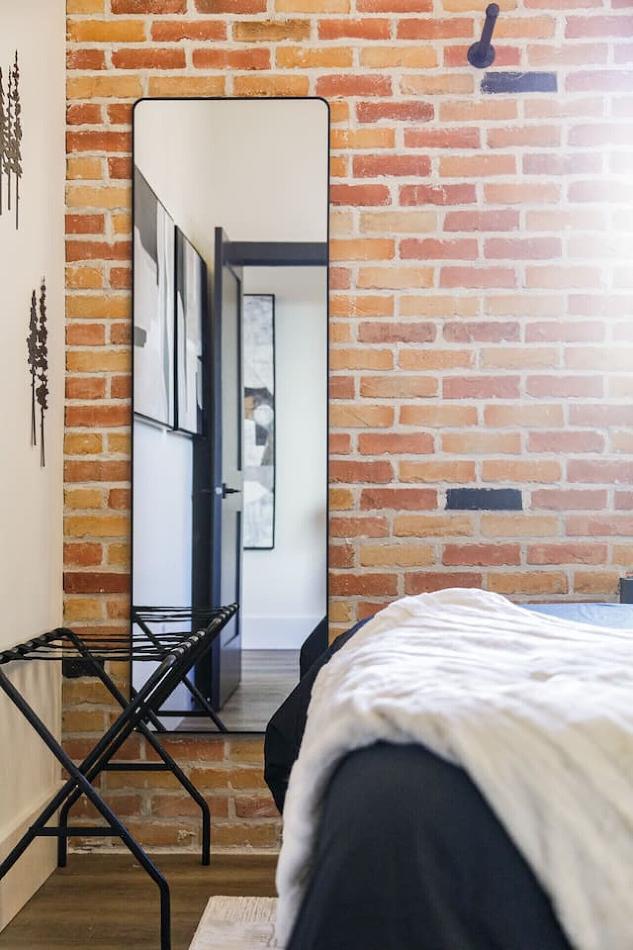 A bedroom with a brick wall, a bed, a desk with a chair, and a large mirror.