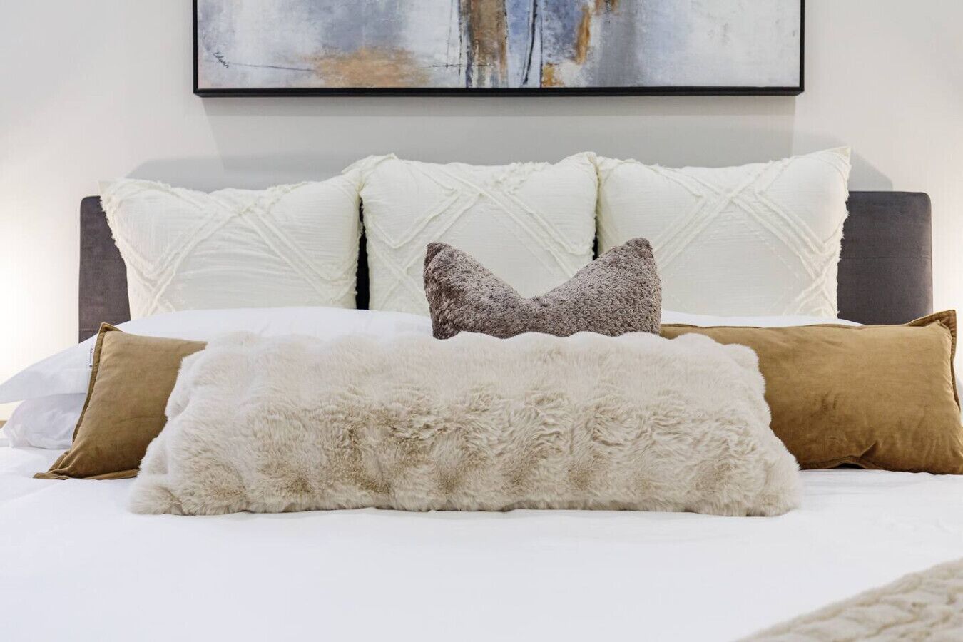 A bed with white and brown pillows, a fluffy white throw, and a dark headboard.